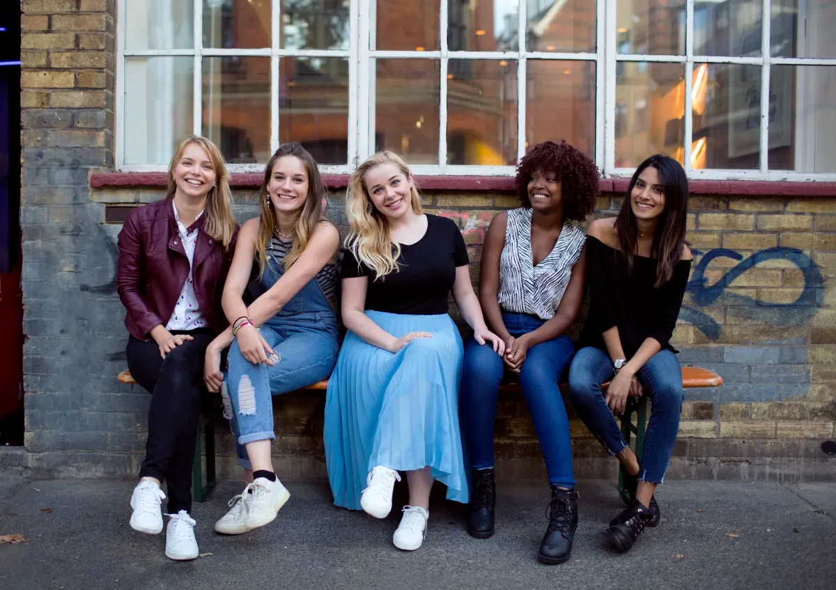 Meninas sentadas em um banco e sorrindo em frente a um prédio.