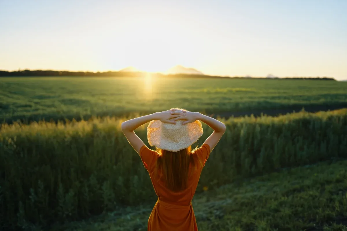Mulher fotografada de costas, observando o pôr do sol em um campo.