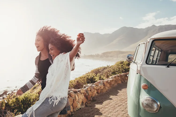 Duas mulheres sorrindo, de pé na beira do mar, ao lado de uma van.
