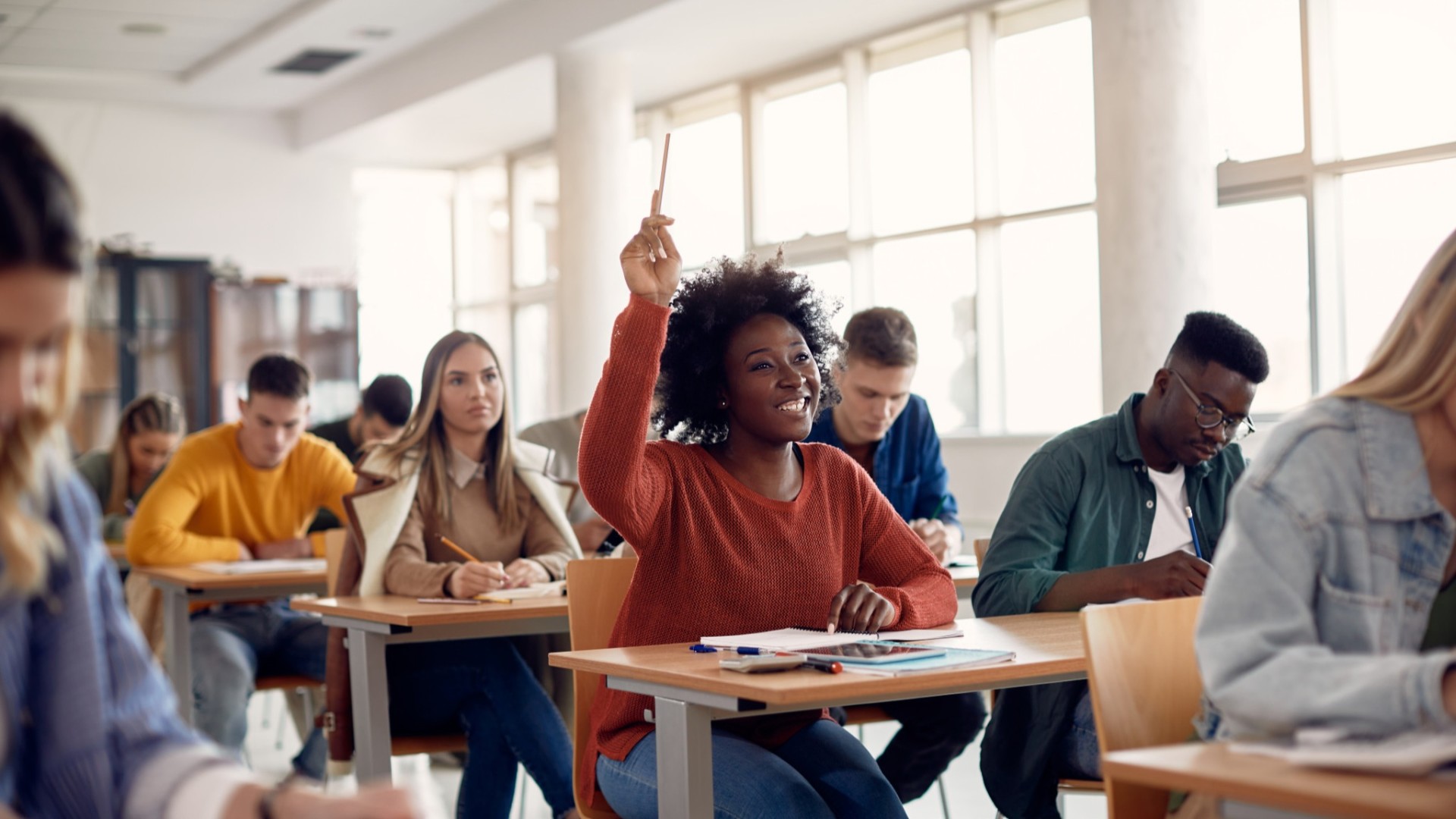 Happy student raising an arm to answer a question while attending class with her university colleagues.