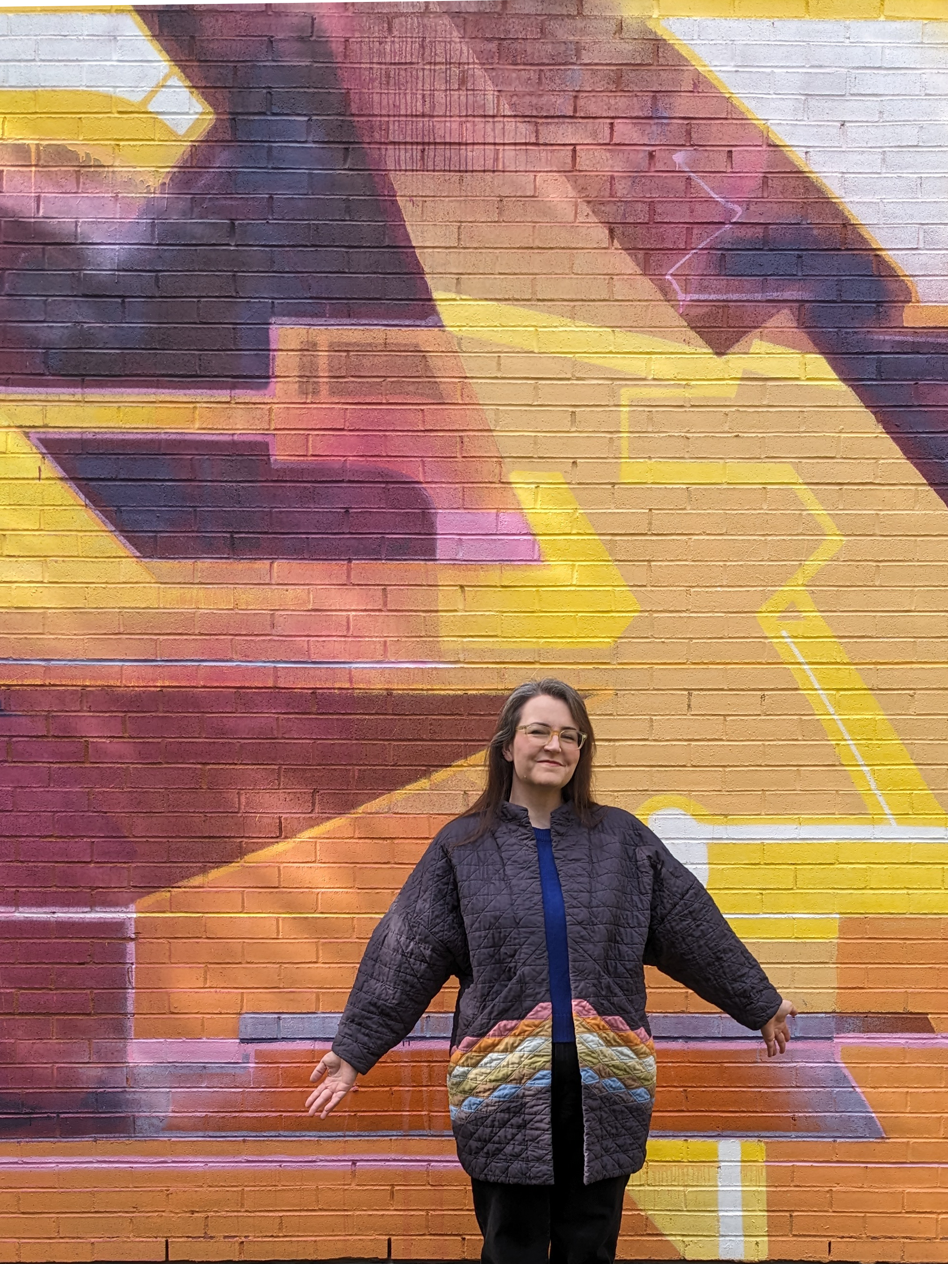 Me standing against a mural in my quilted rainbow coat smiling