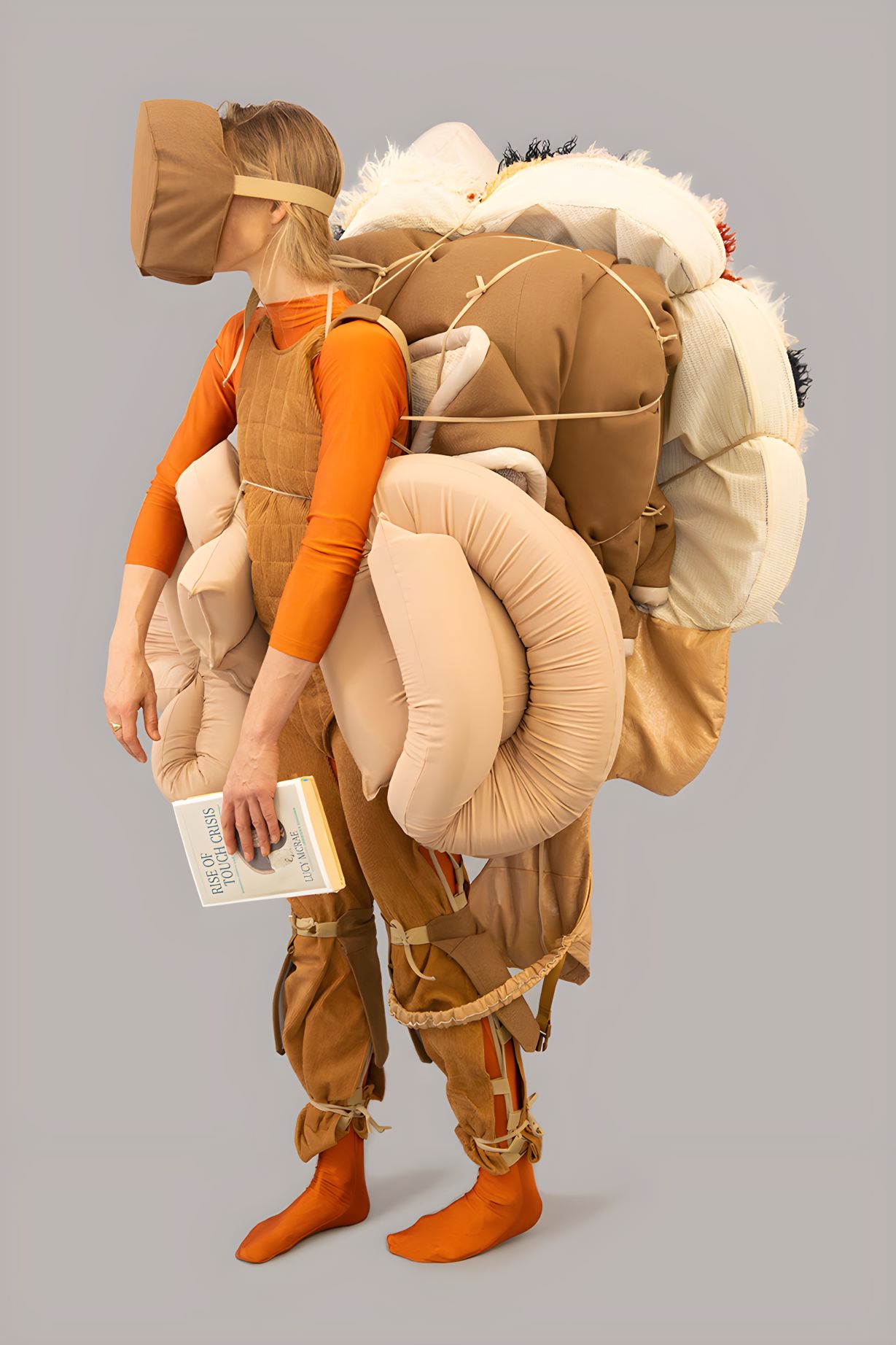 Lucy McCrae performs their work Future Survival Kit - the artist is wearing an orange bodysuit and clad in heavy foam and cloth shinguards, eye-mask and huge backpack. 