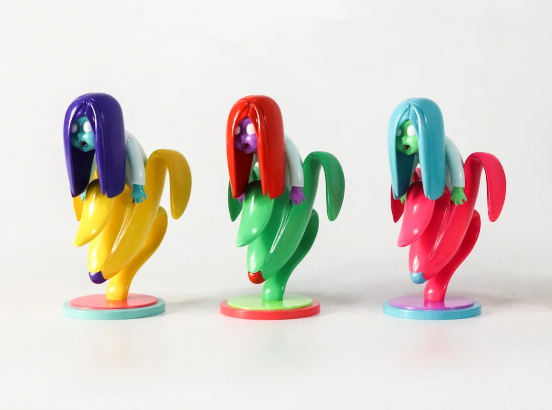 Three banana-shaped character figures with long hair, painted in purple, red and teal colorways on round bases