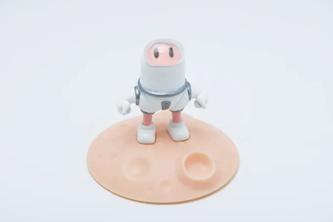 Rounded white astronaut character with a pink face standing on a peach moon base