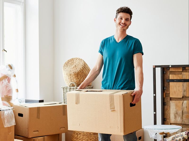 Man in a blue t-shirt stood holding a large moving box.