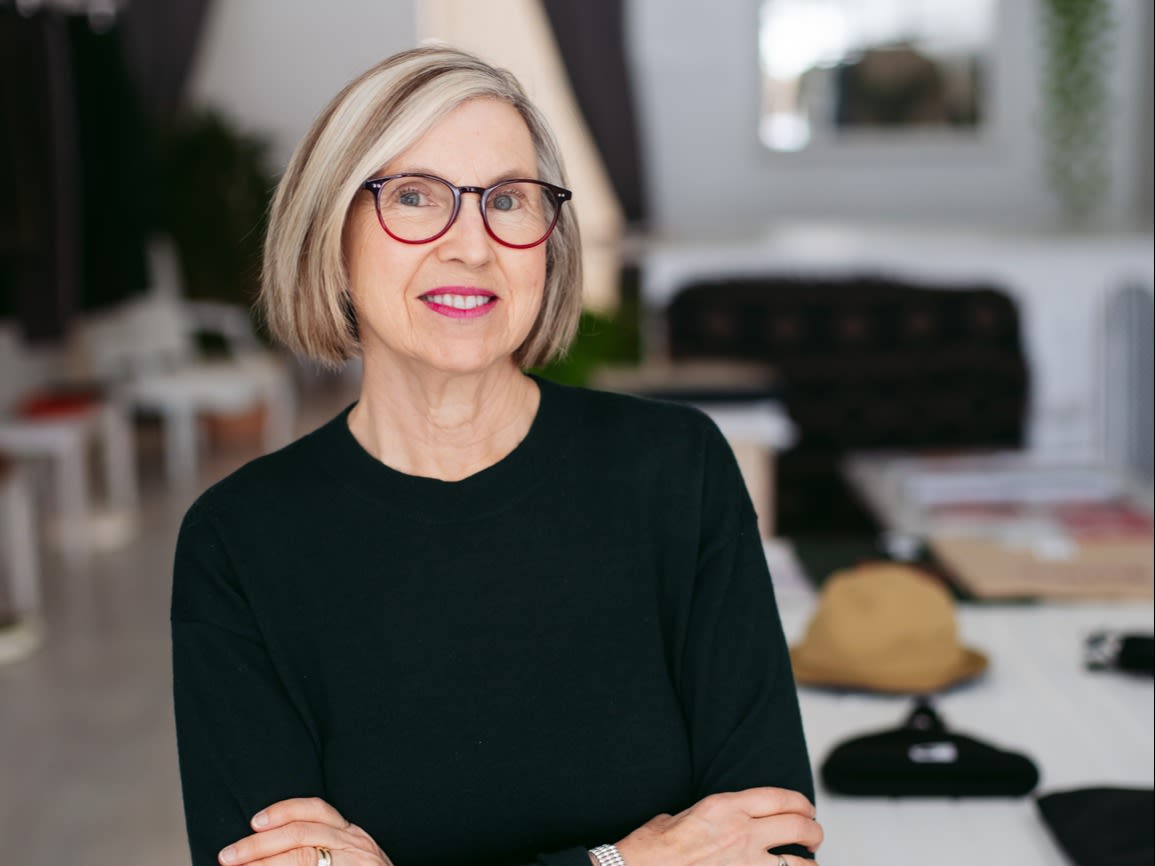 Woman with short hair and glasses, stood with arms folded in front of some desks.