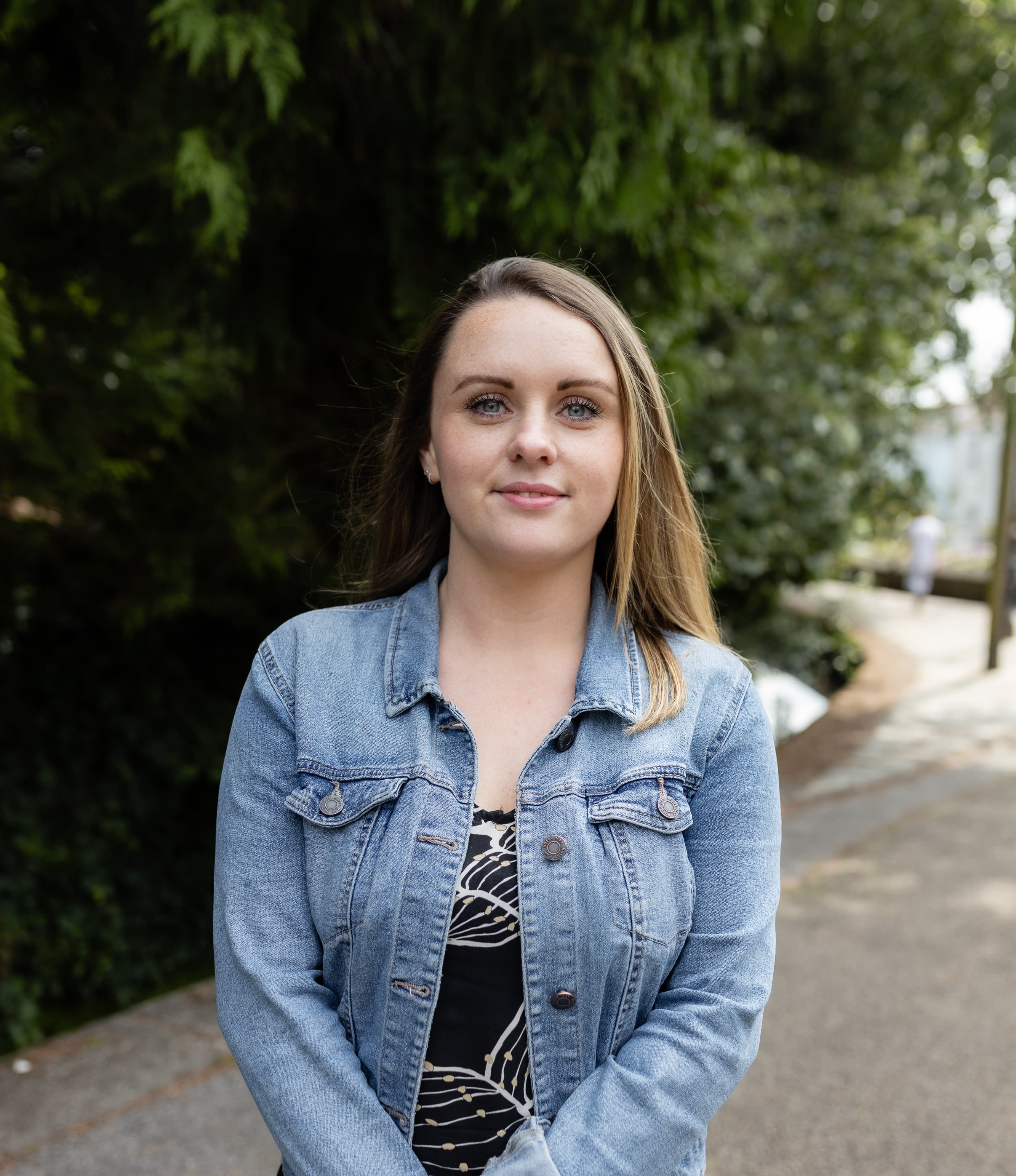 Dark haired woman wearing a blue denim jacket, smiling and standing in front of trees.