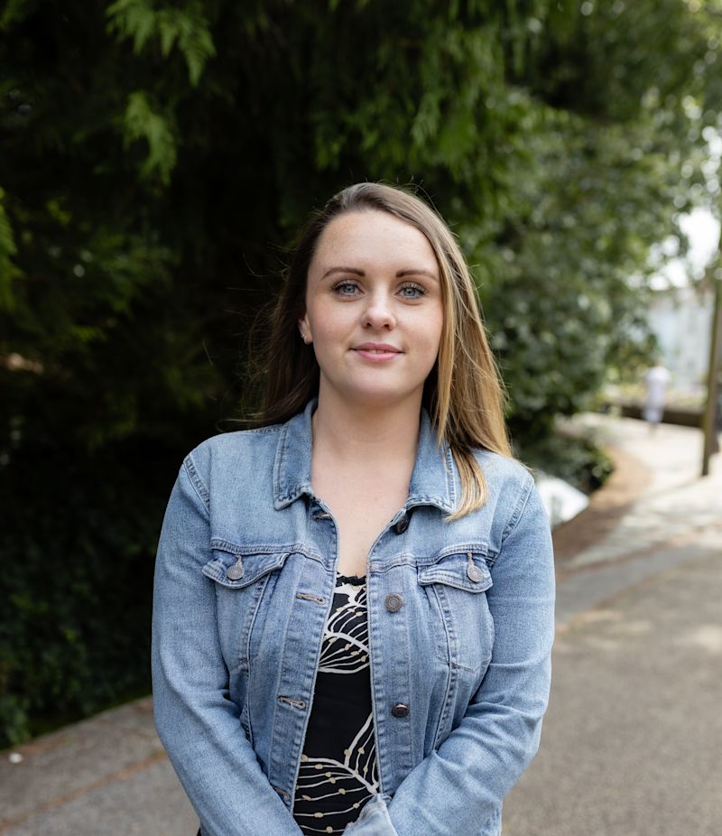 Dark haired woman wearing a blue denim jacket, smiling and standing in front of trees.
