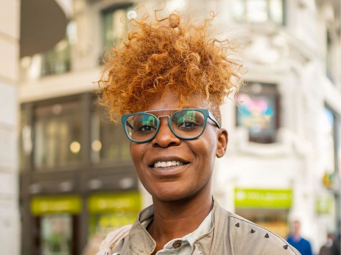 Person with curly hair and green glasses, stood in front of a tall building.