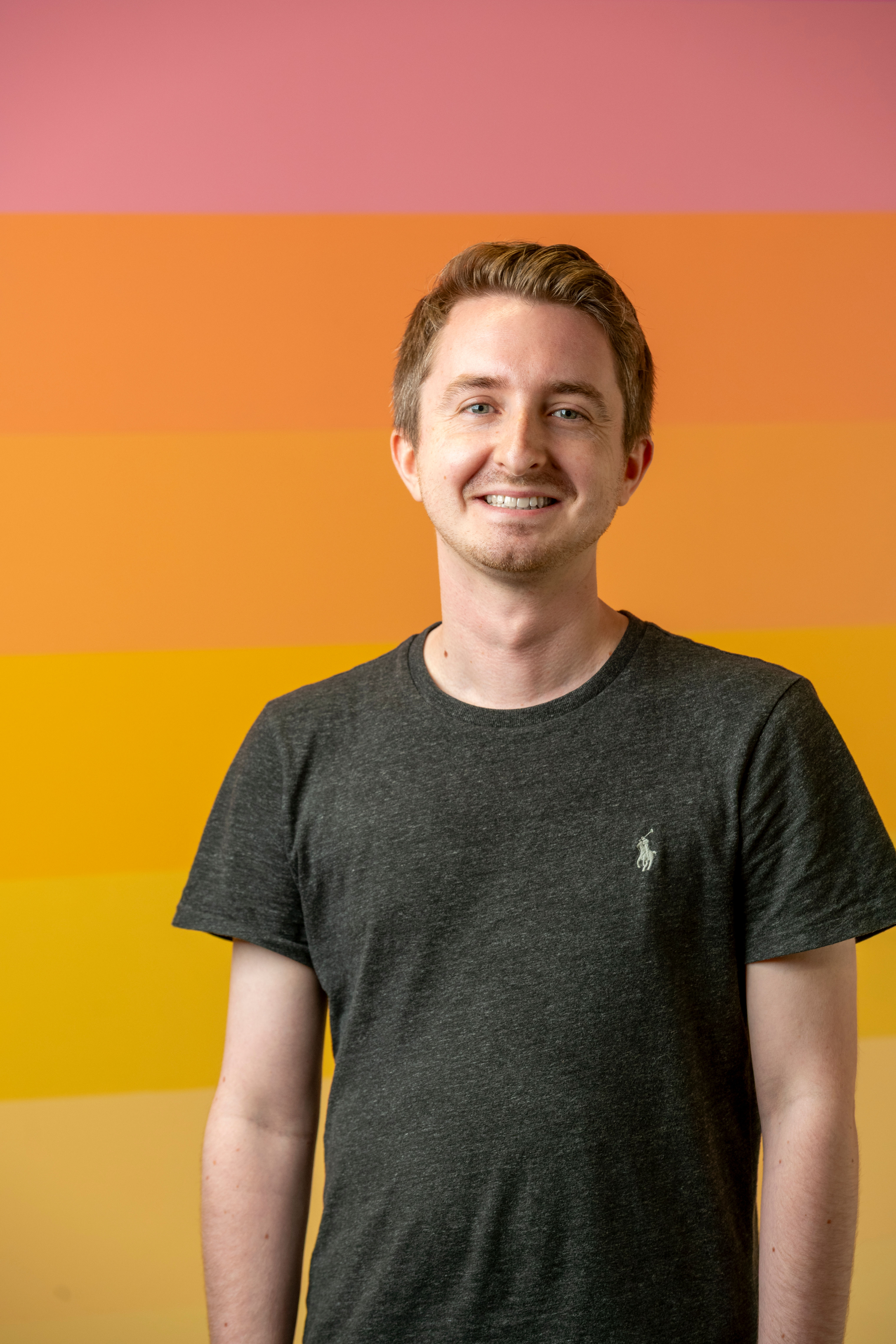 Smiling man wearing a dark grey top in front of an orange gradient background.