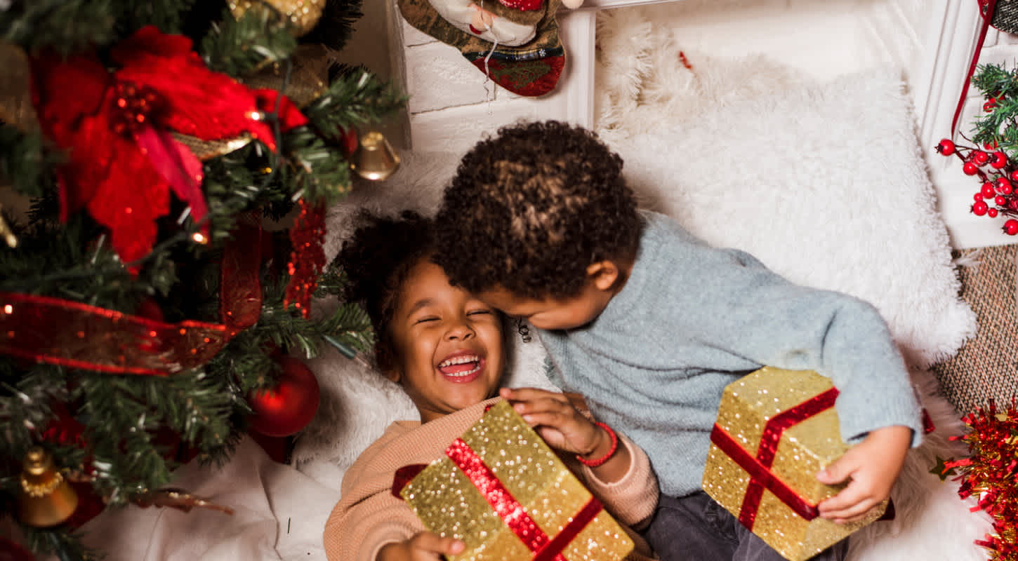 Two young siblings celebrate underneath their Christmas tree while holding gifts in their arms.