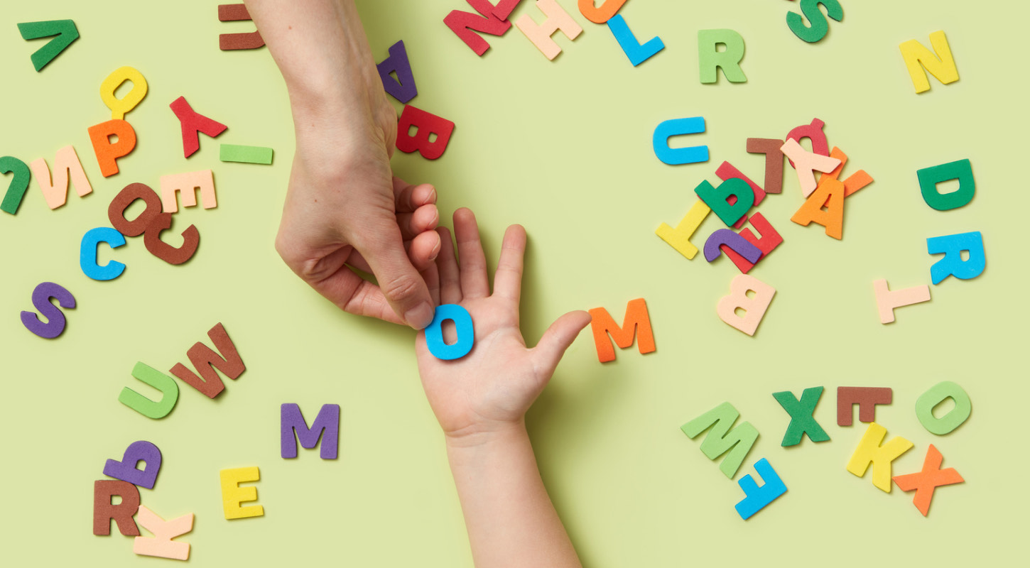 The letter O is picked up from an assortment of alphabet letters scattered on a table and is placed on the palm of an extended hand.
