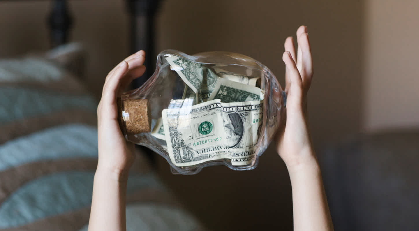 A glass piggy bank is held up in mid-air by the hands of a young child. It contains multiple one-dollar bills and is sealed shut with a cork.