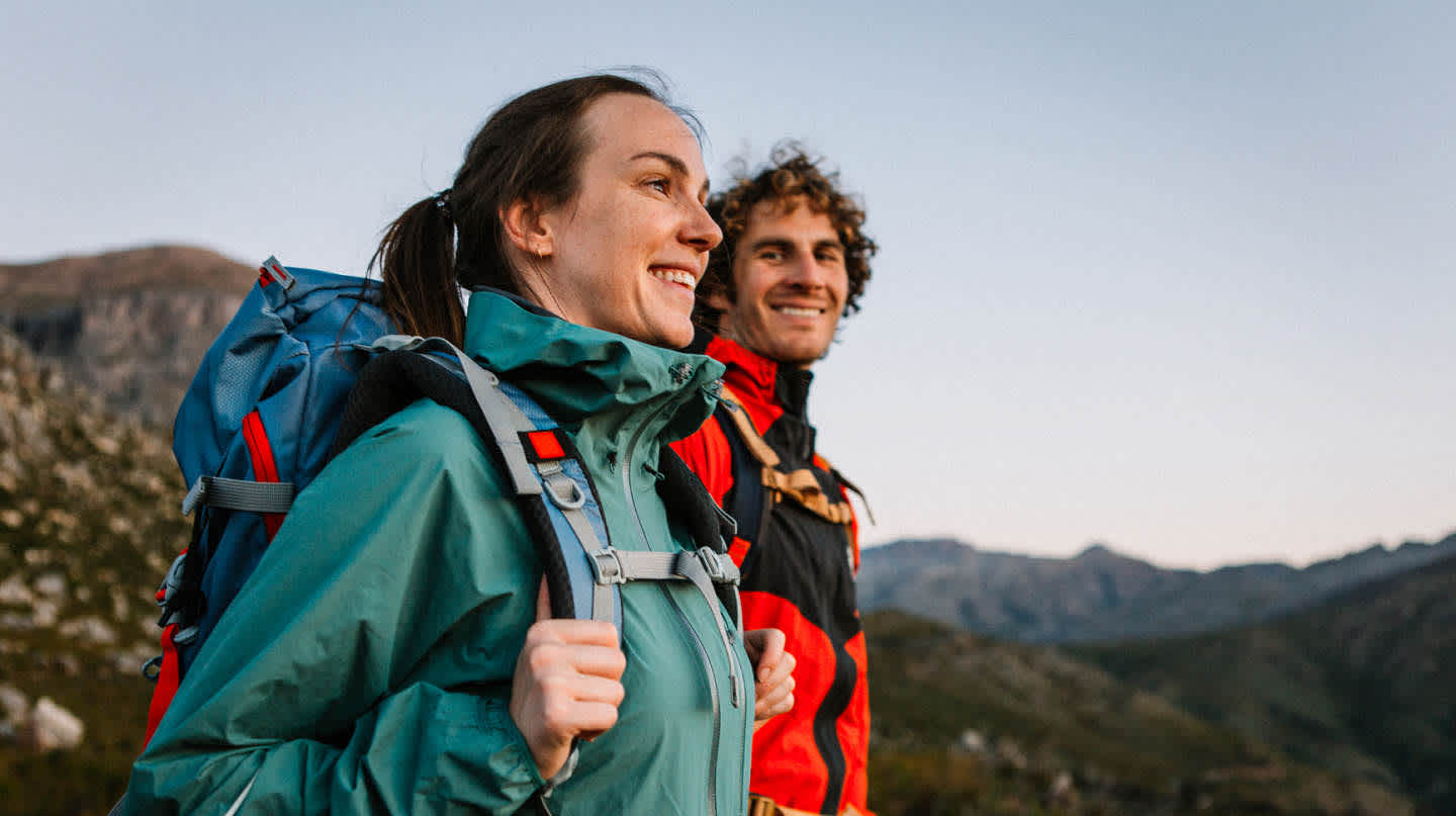 A young couple wearing weatherproof hiking gear gaze out at the scenery from a solitary mountain peak.