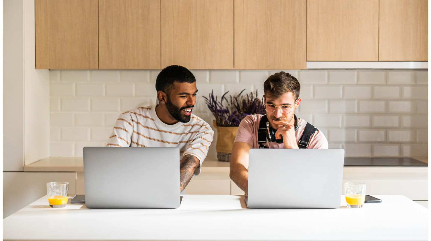 Two young hipster men sitting side by side with open laptops on a kitchen island engaging in conversation over a glass of orange juice. 