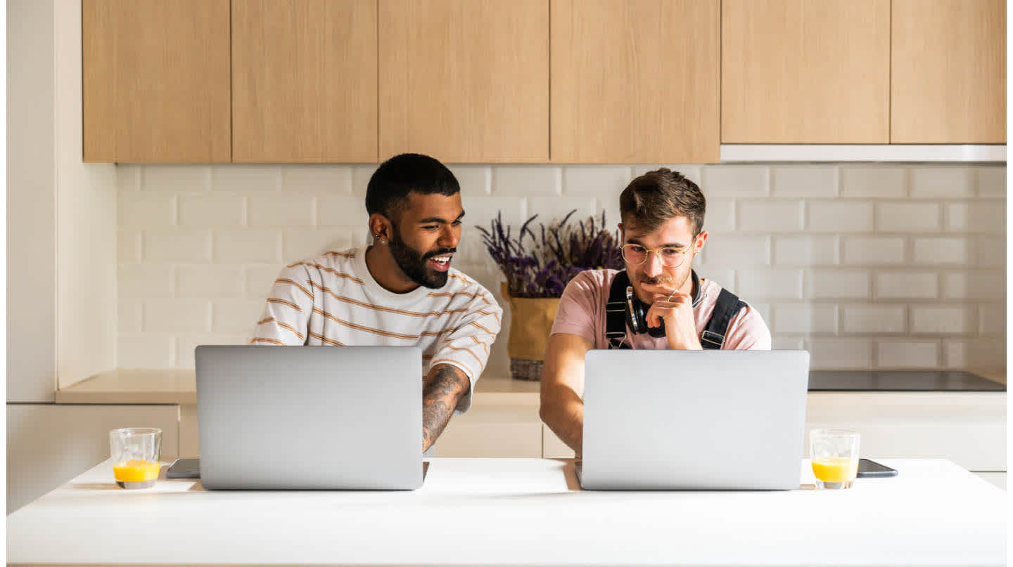 Two young hipster men sitting side by side with open laptops on a kitchen island engaging in conversation over a glass of orange juice.