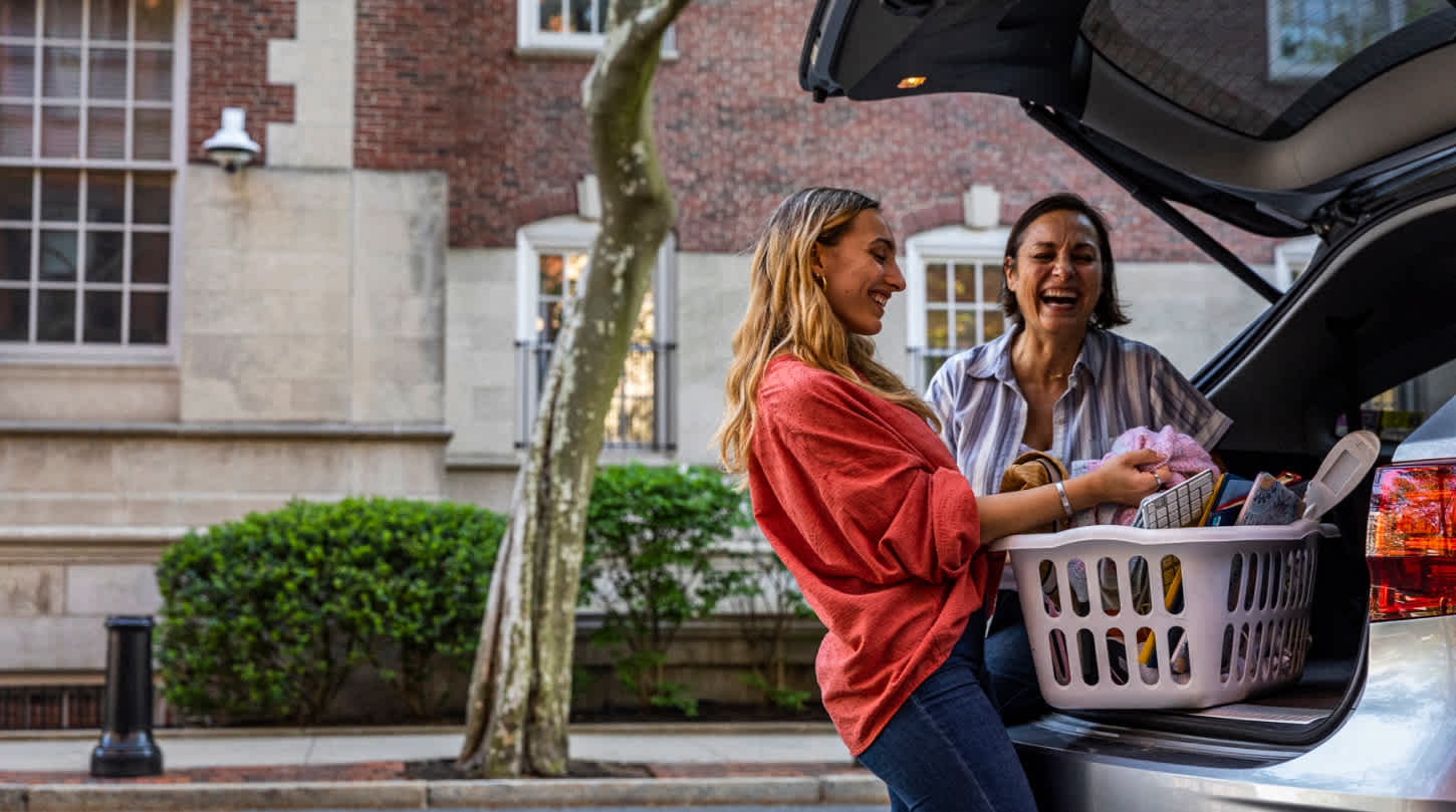 A mother and daughter giggle as they unload a filled laundry basket from the trunk of their hatchback vehicle upon arriving at a college campus for move-in.