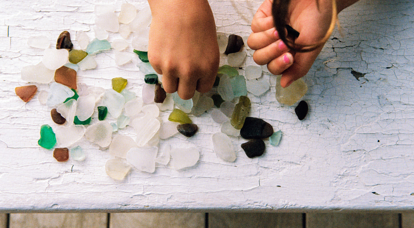A child wearing pink nail polish plays with multicolored gemstones scattered on a table. 