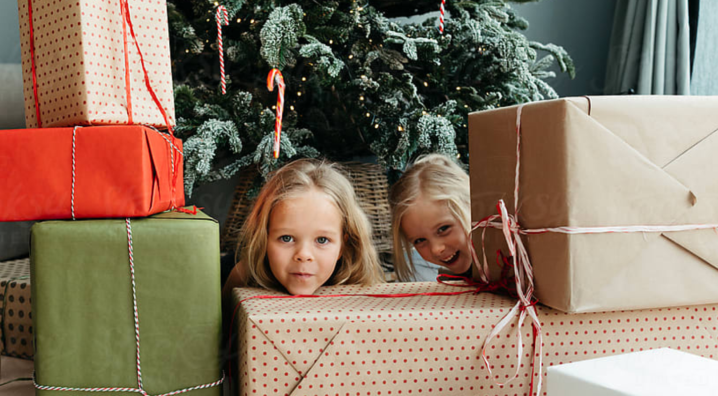 Two young girls pose for a holiday picture underneath a Christmas tree that is filled with presents.