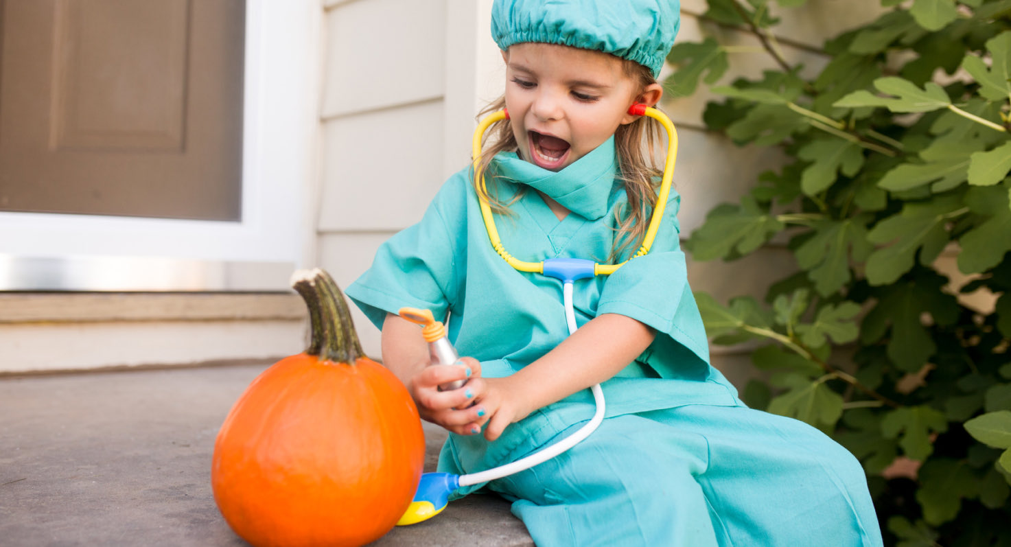 A toddler dressed up as a physician for Halloween wears medical scrubs and listens through a stethoscope while sitting on a stoop alongside a pumpkin.