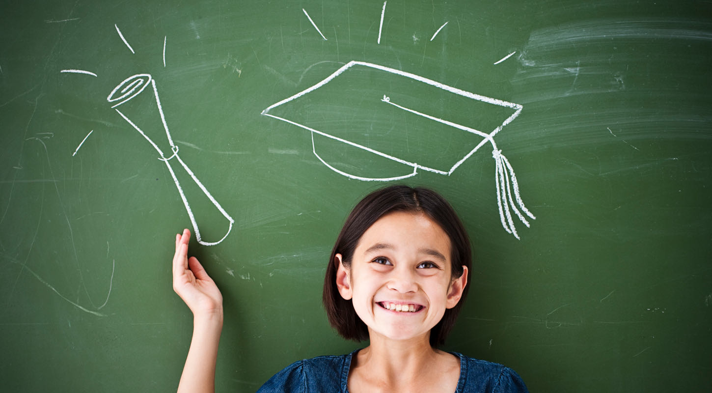 A bright eyed school age girl stands in front of a chalk board. She holds a drawn diploma and wears a drawn graduation cap. 