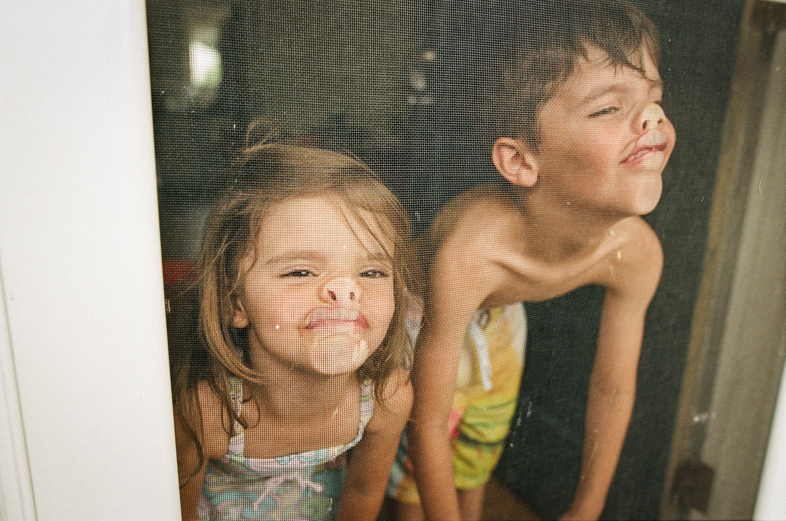 A young girl and boy with "rag doll" posture pressing their faces against a window screen, making their noses and mouths smoosh up - parents are more burnt out