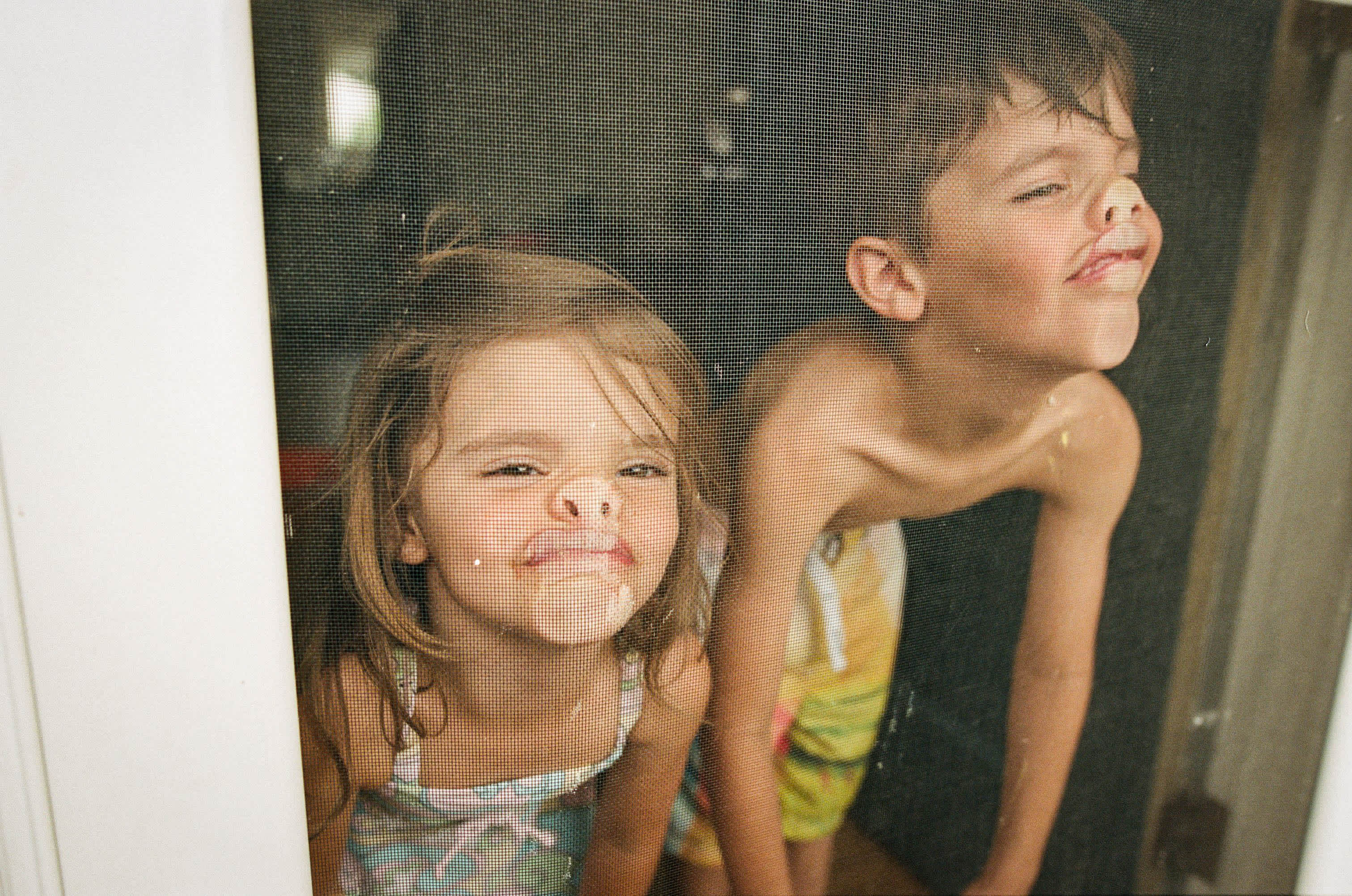 A young girl and boy with "rag doll" posture pressing their faces against a window screen, making their noses and mouths smoosh up - parents are more burnt out