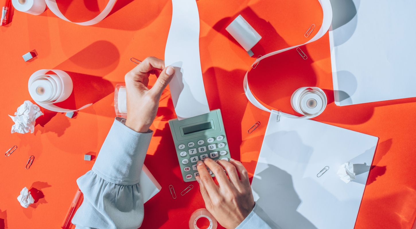 A person operates a printing calculator, fingers swiftly tapping the keys, surrounded by scattered papers atop a cluttered desk.