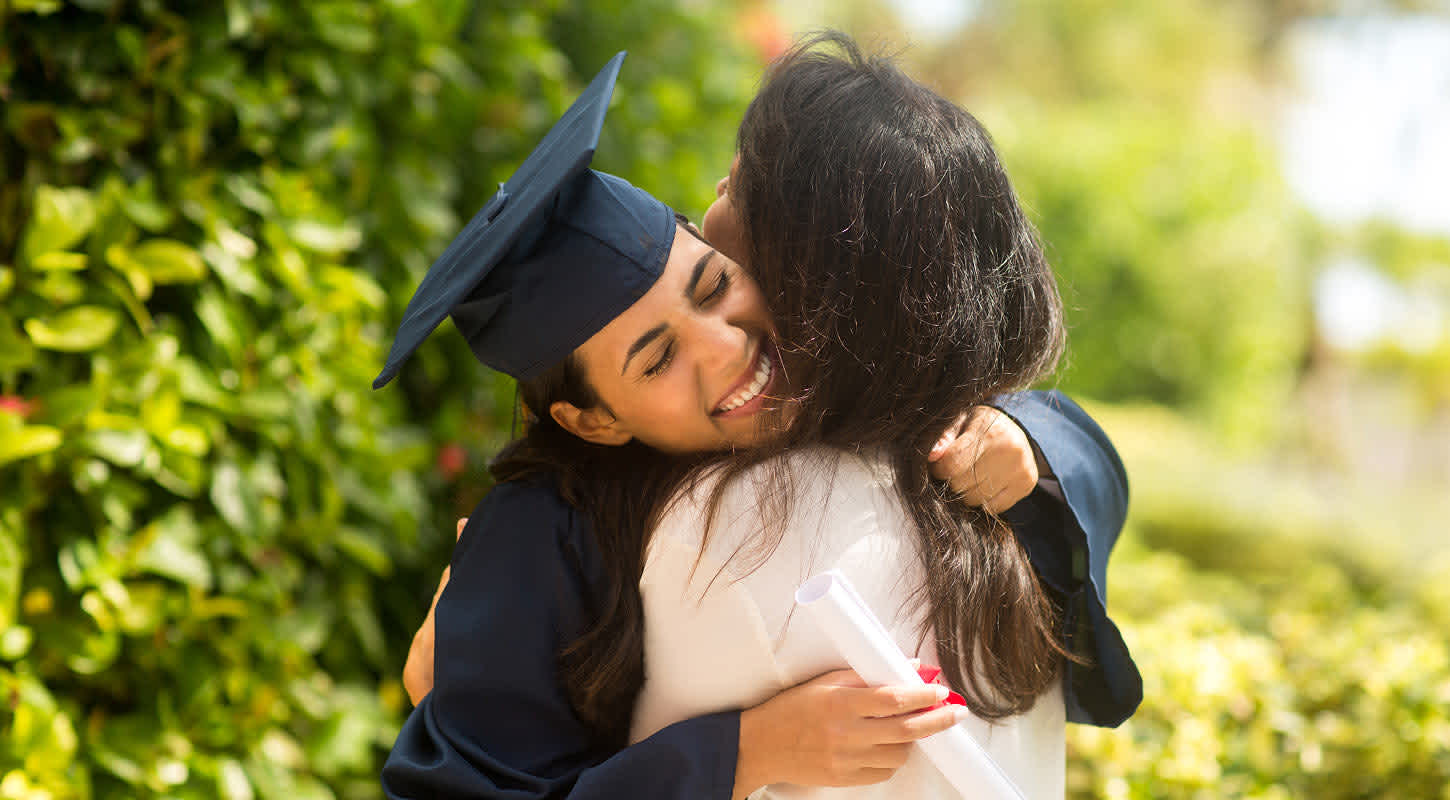 A young graduate in a cap and gown embraces an older woman, sharing a joyful hug.