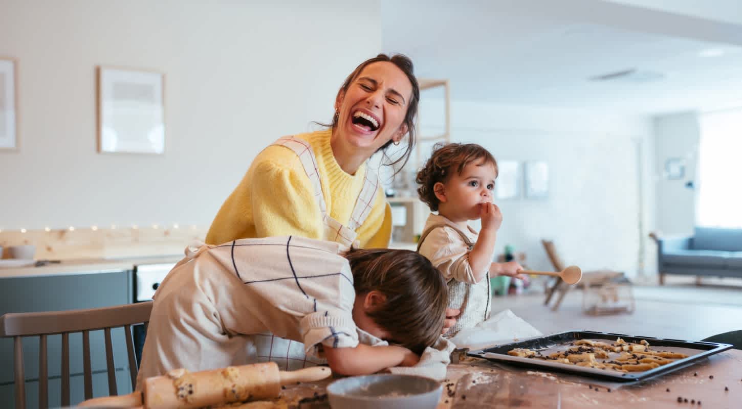 A cheerful young mother laughs hysterically while standing in front of a cookie-filled baking sheet on a countertop covered in chocolate chips. She is joined by her two curious toddler-aged boys, all enjoying the moment together.