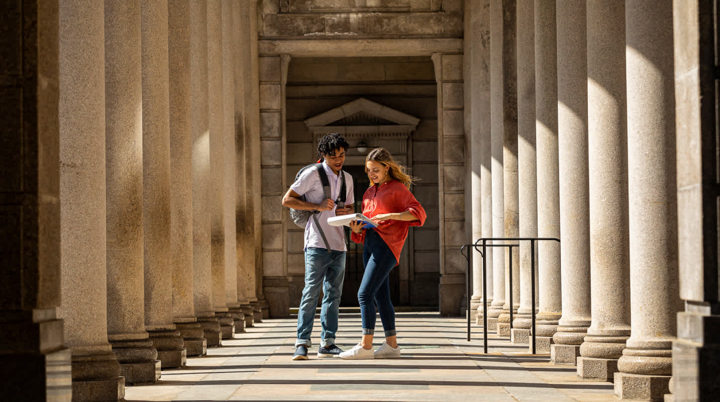 Two classmates closely review a document inside of an open binder while standing in the middle of a pillared archway.
