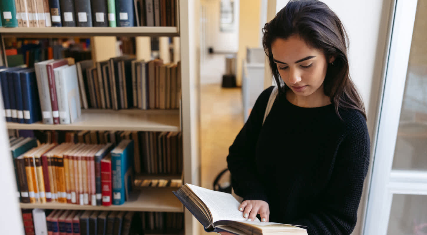 A young woman stands in the aisle of a library filled with books, engrossed in an open book she holds in her hands.