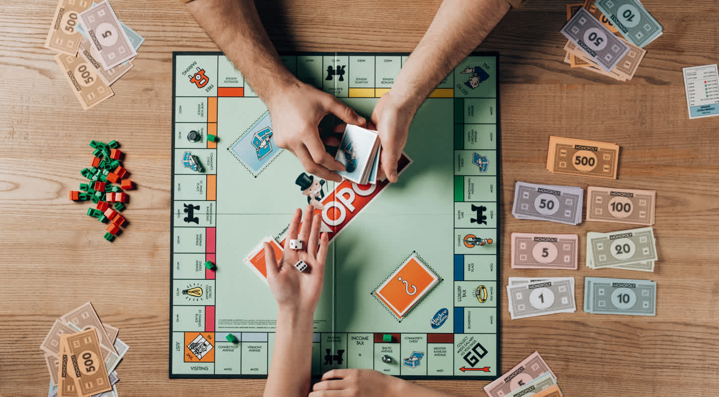 A game of monopoly is underway. Two players with stacks of monopoly money are shown rolling the dice and shuffling cards.