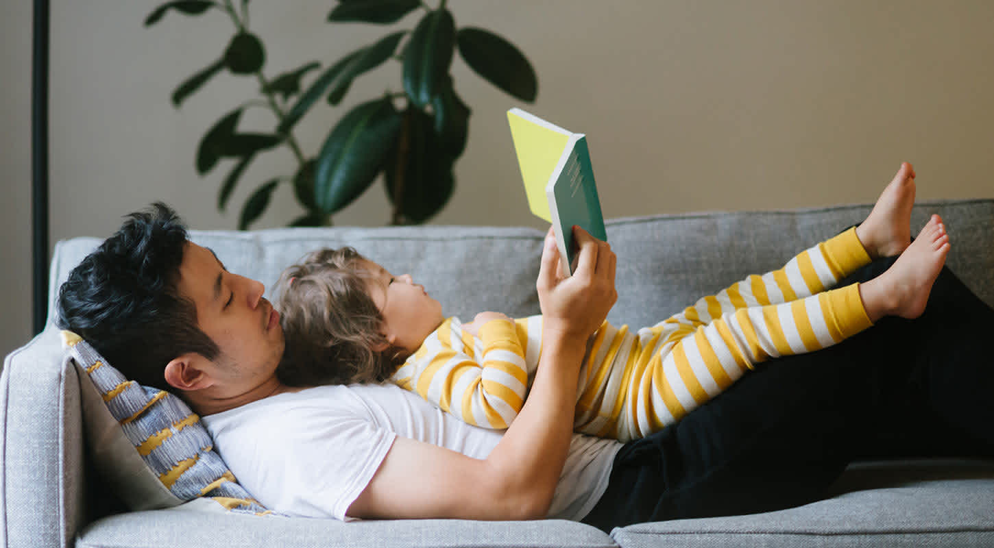 A young father, stretched out on a couch, reads a book to his toddler lying on his belly.