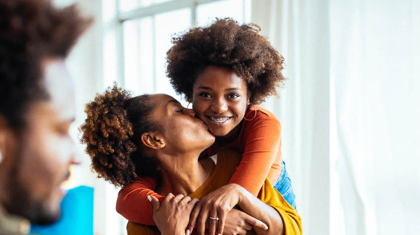 A candid mother-daughter embrace. A young daughter wraps her arms around her mother who is sitting down. She smiles as she receives a kiss on the cheek from her mother. Both sport an afro, as does the father in the foreground.