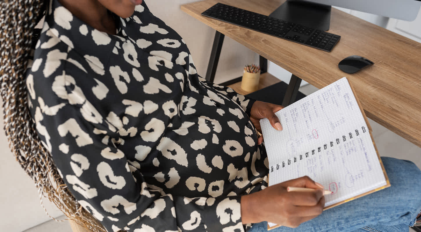 An expecting mother sits at her desk with an open journal and pencil in hand.