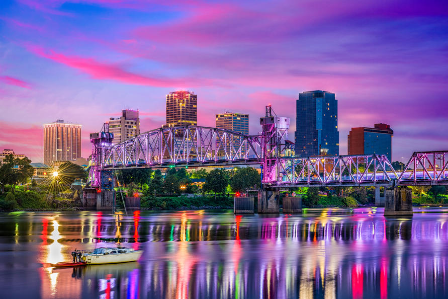 10 Best Cities in America for Single Parents - Waterfront view of downtown Little Rock, AR skyline during sunset with a boat traveling along the bank of the Arkansas River in the foreground.