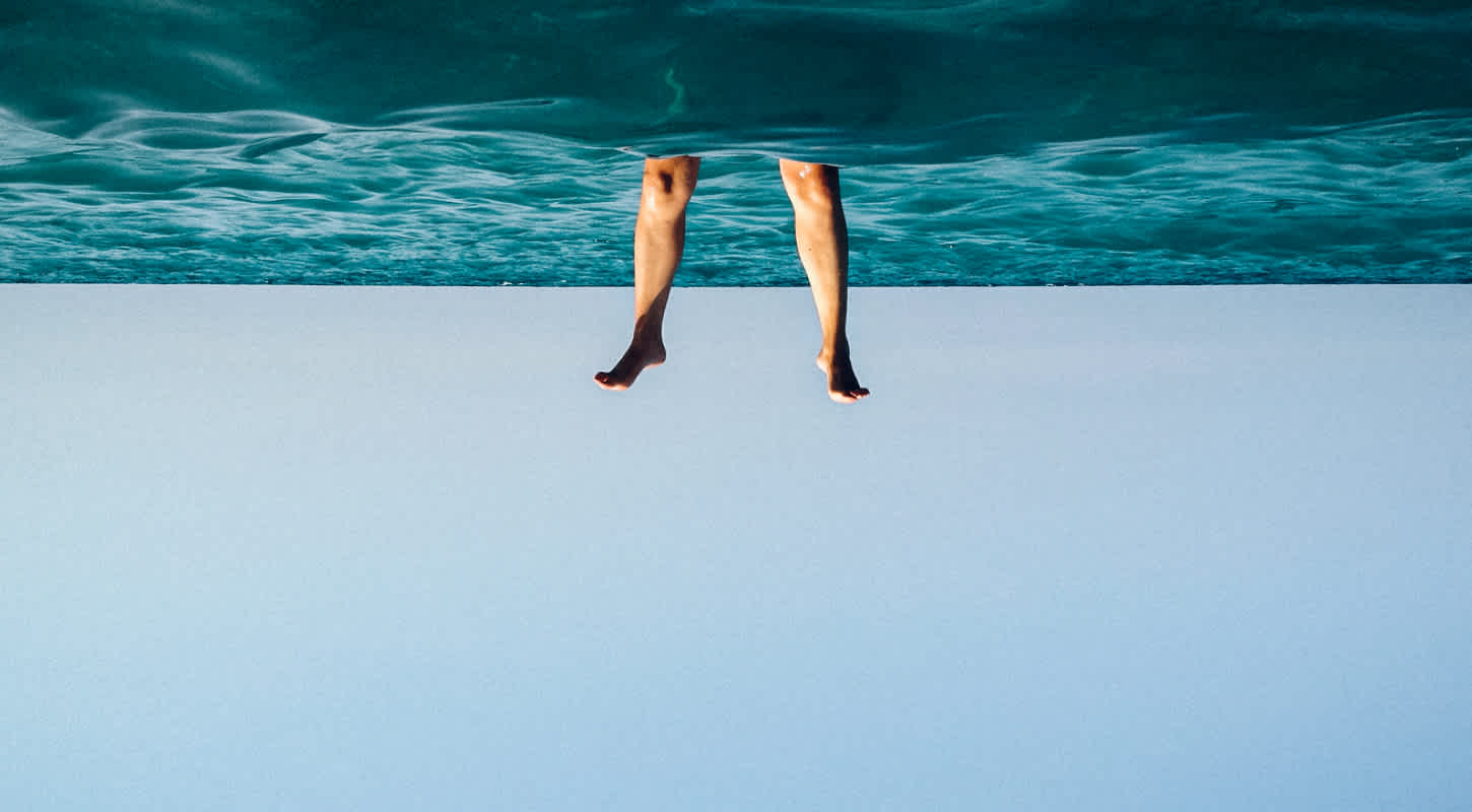 An inverted seascape depicts a person submerged head first up to their knees in a vast body of water.