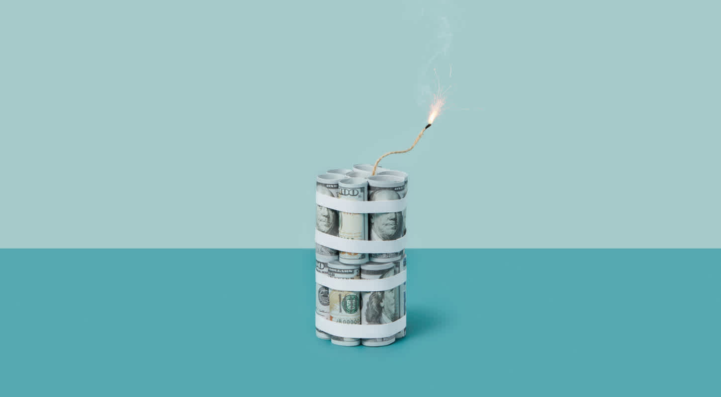 Multiple one-hundred dollar bills are rolled up and fastened in two stacks to resemble a dynamite stick with a lit fuse.