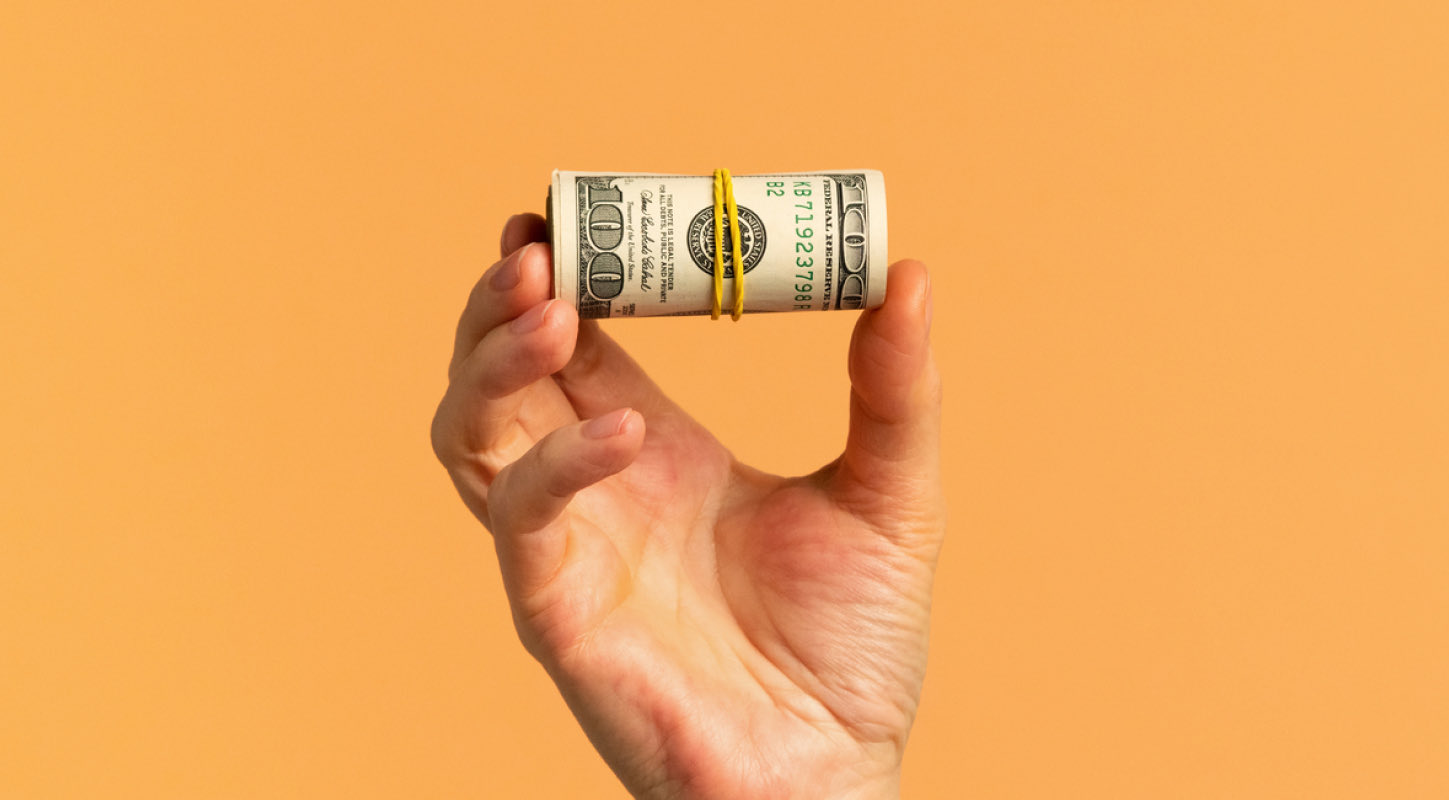A hand holds a rubber-banded wad of one hundred dollar bills in mid-air. 