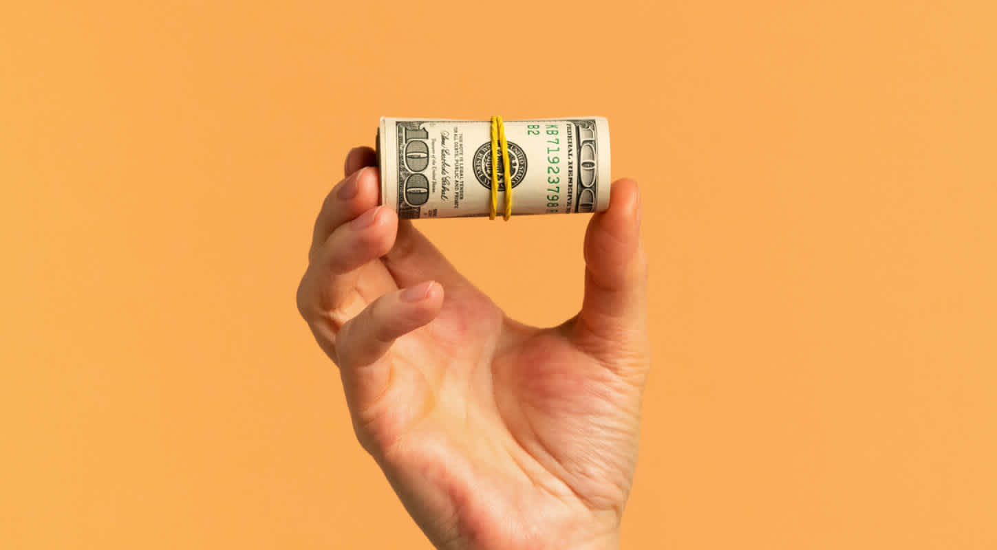 A hand holds a rubber-banded wad of one hundred dollar bills in mid-air.
