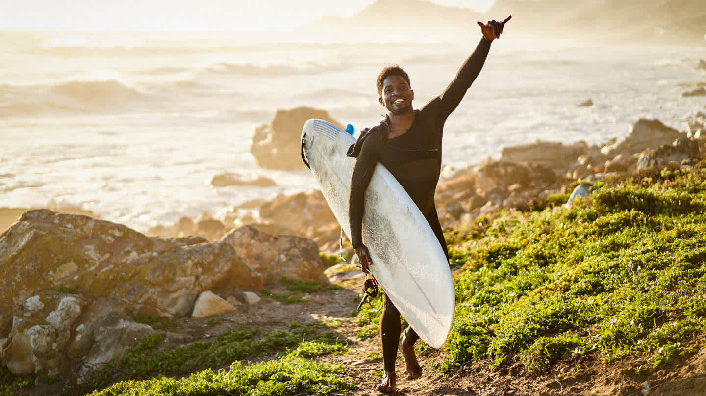 A surfer carrying a longboard and wearing a seat suit makes a pura vida hand gesture while walking away from the surf.