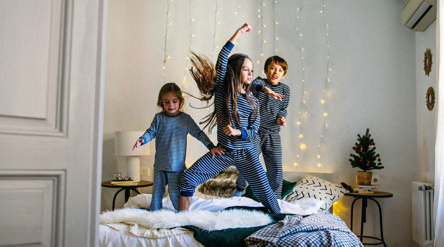 Three children ranging from toddlers to tweens jump playfully on a bed while in their pajamas. 