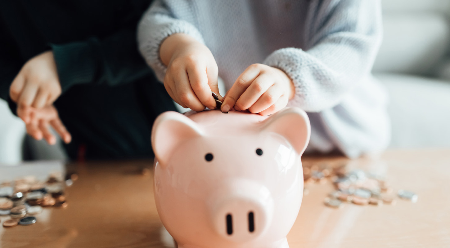 Two children standing in front of a table with a pile of coins begin to stuff them inside a pink piggy bank. 