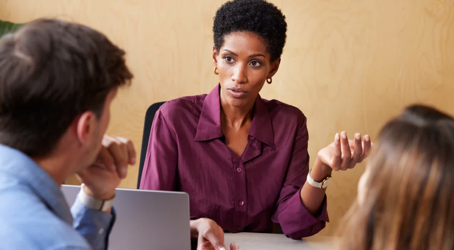 A businesswoman leans forward at a meeting table, gesturing confidently with her hands as she engages with colleagues across from her.