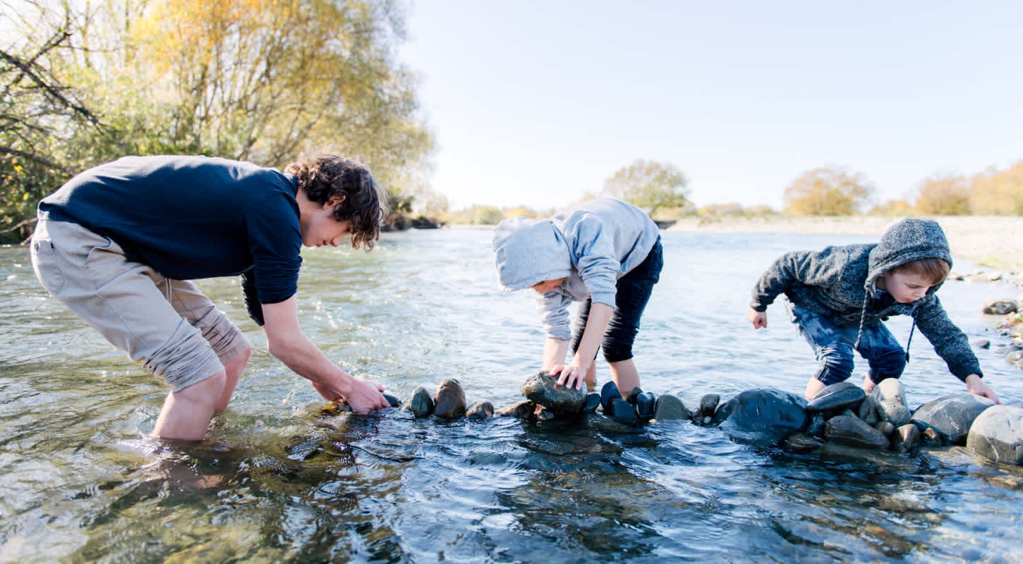 Three children submerged ankle deep in water play with rocks on the bank of a creek.