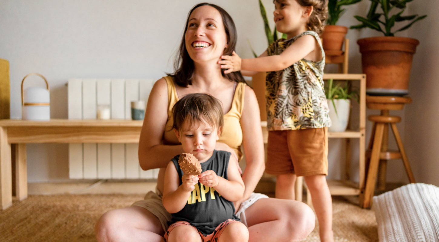 A young mom sits crossed-legged on a rug. One of her toddlers sits in her lap, while the other stands beside her and plays with her hair. 