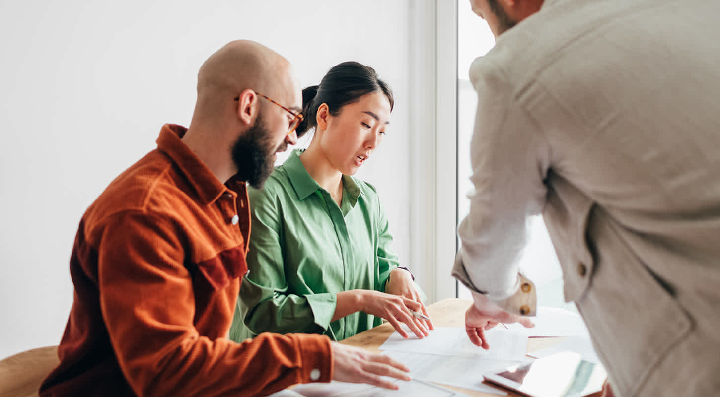 A couple sits at a table reviewing a contract while another person, likely a professional or advisor, gestures over the documents to provide guidance.