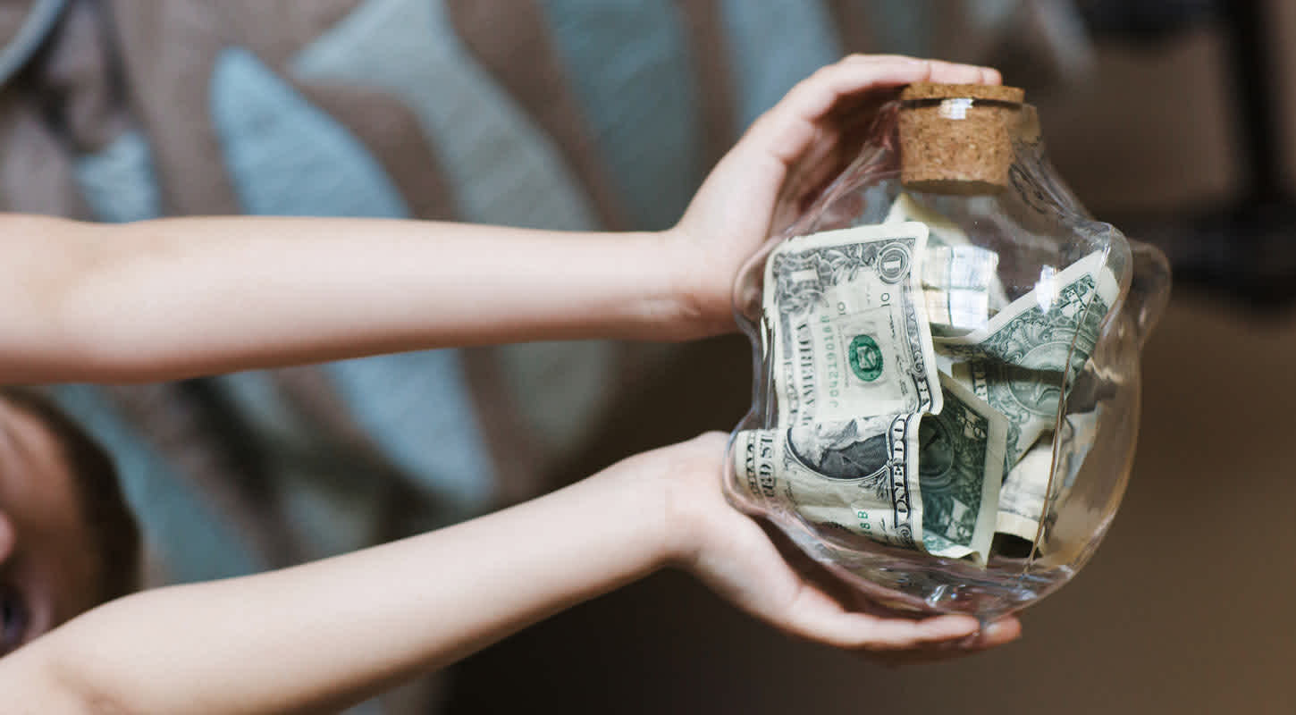 Two hands hold a glass piggy bank with saved dollar bills in mid-air.