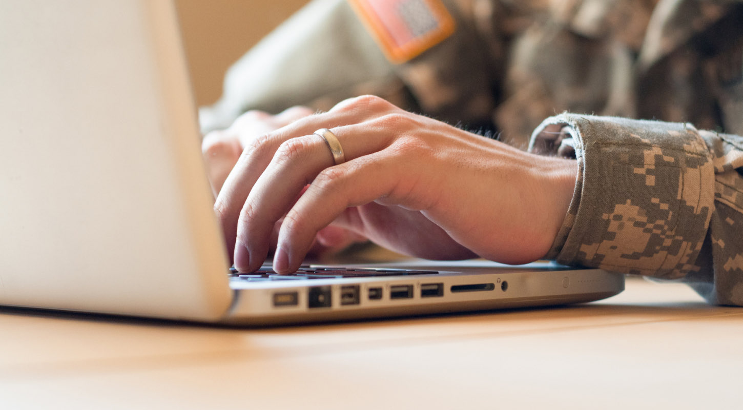 A service member in their military uniform wears a wedding band on their ring finger while typing on a laptop.
